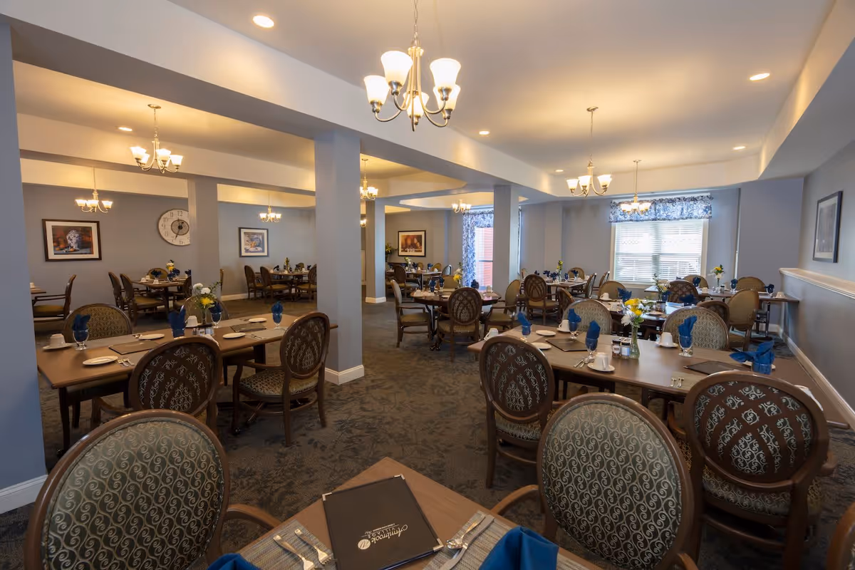 A spacious dining room with multiple tables and chairs arranged neatly. Each table is set with plates, cups, silverware, and blue napkins. The room has soft lighting from chandeliers hanging from the ceiling, light blue walls, carpeted floors, and windows with blue patterned curtains allowing natural light to enter.