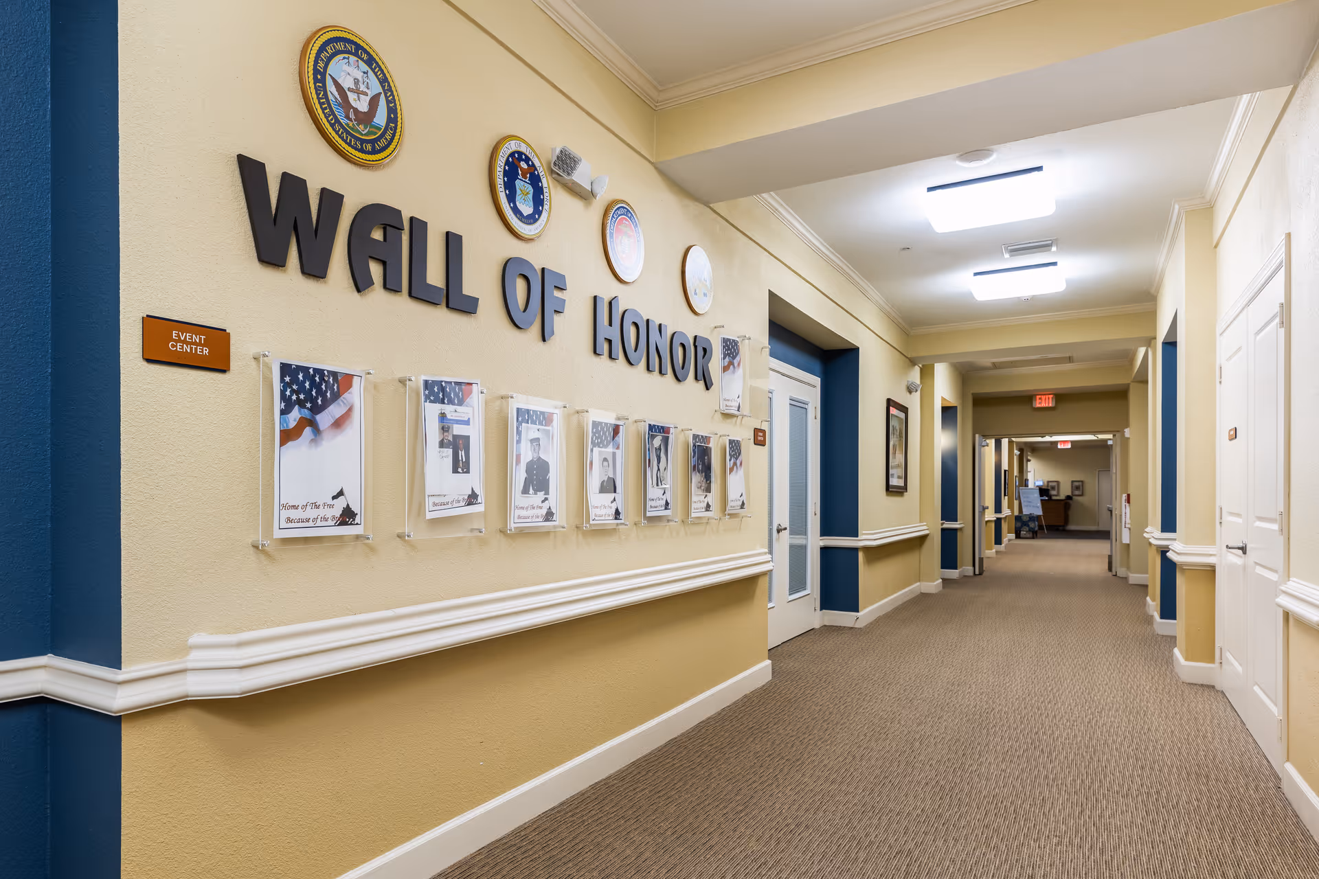 Long indoor hallway in a senior living facility with a 'Wall of Honor' display and framed photos along the left wall.