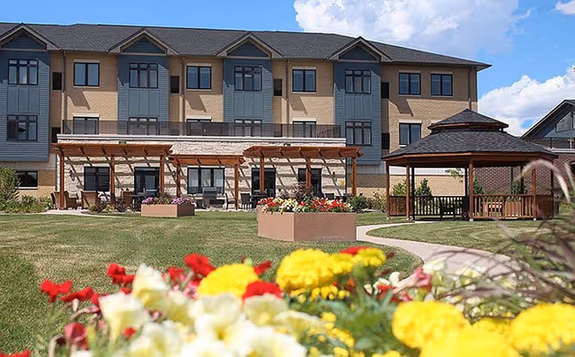 Exterior view of a multi-story senior living facility with a well-maintained garden in the foreground featuring colorful flowers. The building has multiple windows and a covered patio area with seating. There is also a wooden gazebo on the right side of the image under a partly cloudy sky.