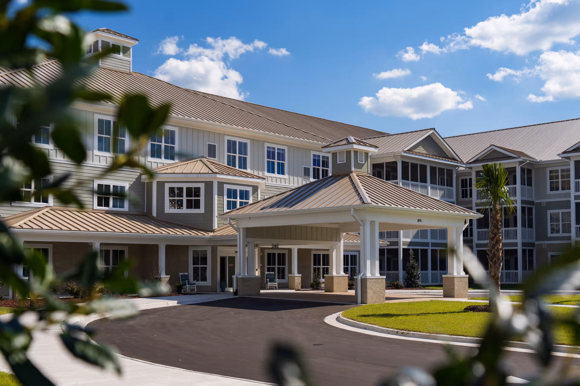Exterior view of a large senior living facility building with a covered entrance driveway, multiple windows, and a well-maintained lawn under a blue sky with scattered clouds. Some green foliage is visible in the foreground.