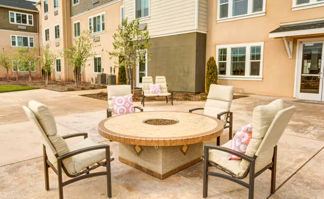 Outdoor courtyard patio at Carmel Village with cushioned chairs arranged around a round stone fire pit in front of the building.