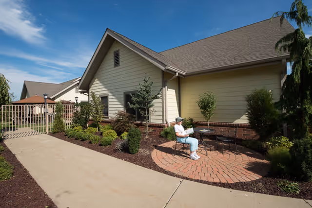 A small circular brick patio beside a pale yellow building with landscaping, where a seated person reads at a table under a blue sky.