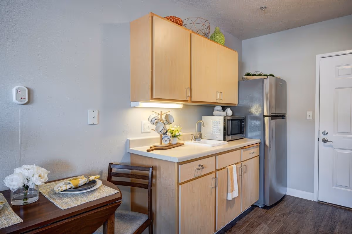 A small kitchen area with light wood cabinets, a white countertop with a sink, a microwave, and a stainless steel refrigerator. A wooden dining table with a chair is partially visible, set with a plate, napkin, and flowers in a vase. The walls are painted light gray, and there is a white door to the right.