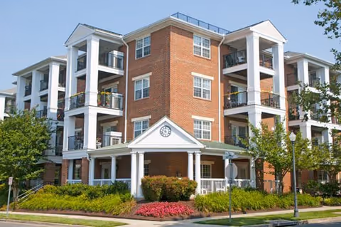 Exterior view of a multi-story brick senior living building with white columns and balconies, surrounded by landscaped greenery and trees under a clear blue sky.