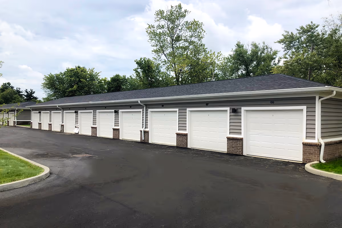 A row of attached garages with white doors and gray siding, situated along a paved driveway with green grass and trees in the background under a cloudy sky.