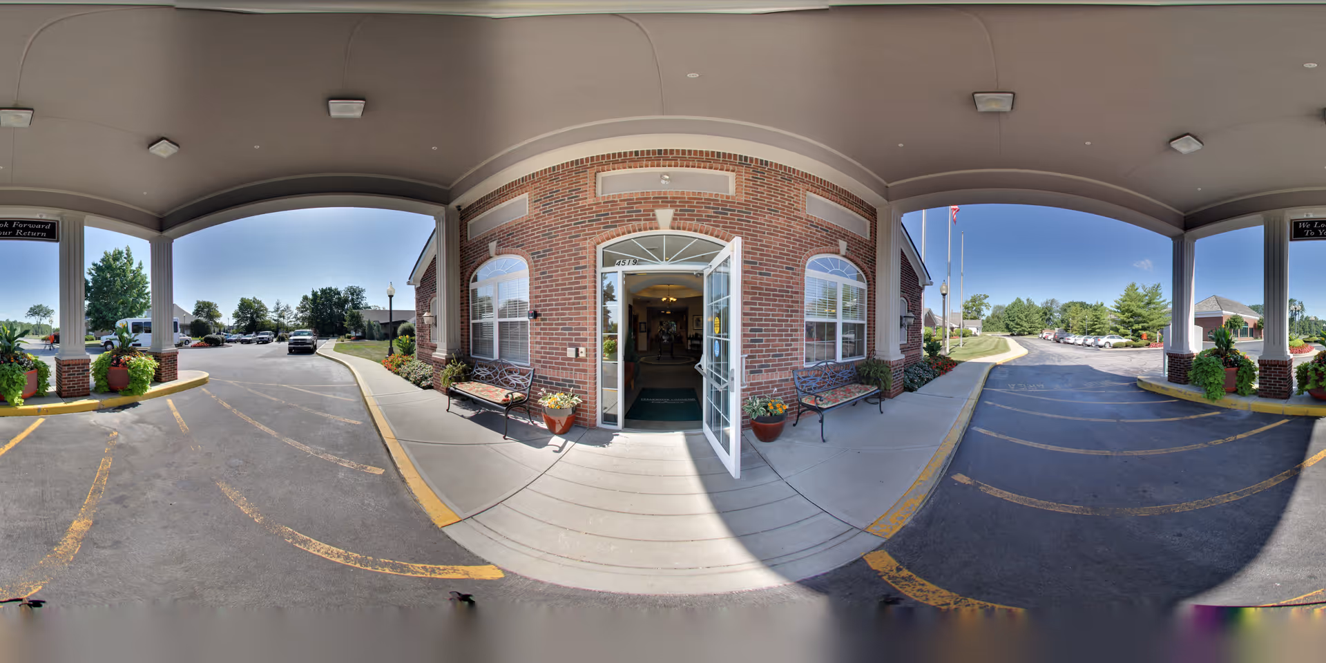 Wide-angle view of the entrance to Five Star Residences of Clearwater, showing a covered driveway with brick pillars and large planters with flowers. The main entrance door is open, flanked by two benches and windows on either side. The parking lot and greenery are visible in the background under a clear blue sky.