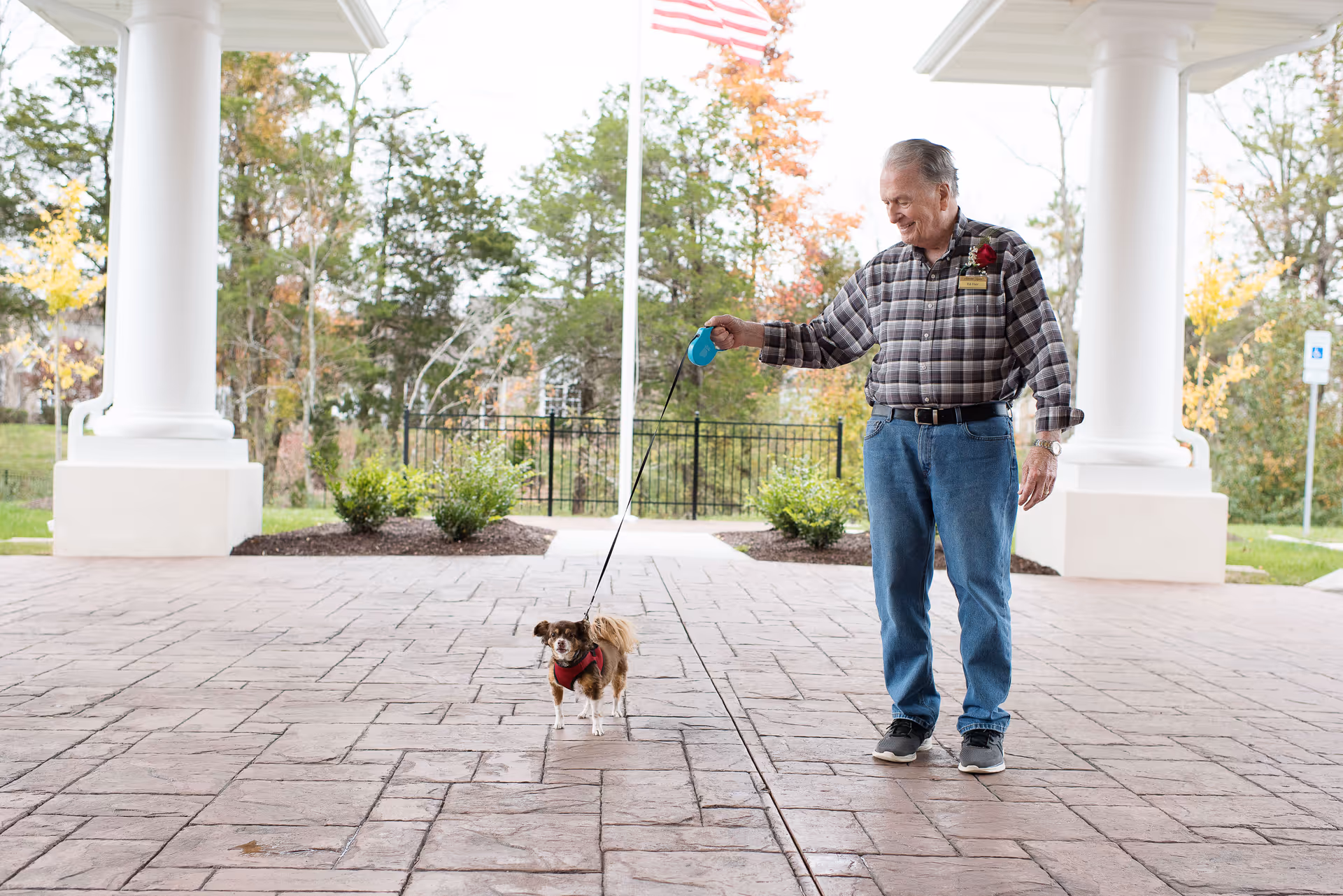 An elderly man stands under a covered building entrance holding a small dog on a leash.