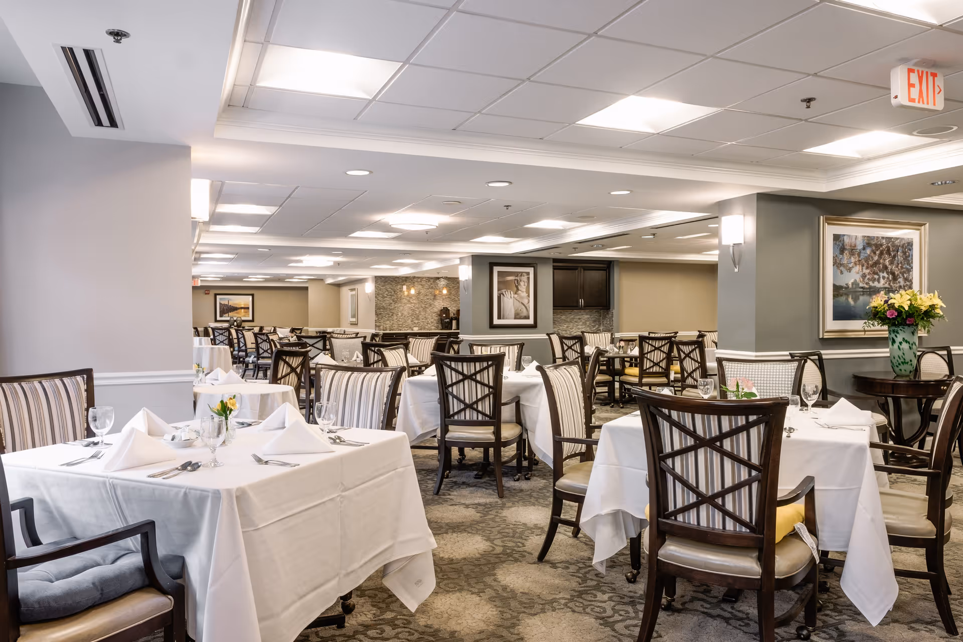 A spacious, well-lit dining room at Thomas House Retirement Community with multiple tables covered in white tablecloths, set with folded napkins, glassware, and silverware. The room features cushioned wooden chairs, framed artwork on the walls, a floral arrangement on a side table, and a carpeted floor with a patterned design.