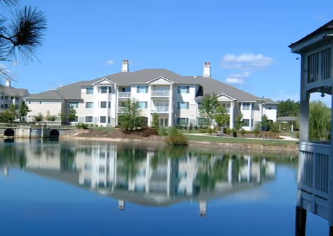 Three-story white senior living building reflected in a calm pond, with a gazebo visible at right.
