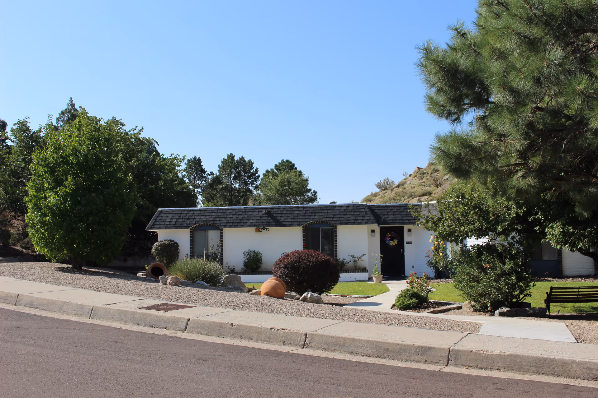 Single-story white residential building with a dark shingled roof, surrounded by green trees, bushes, and a well-maintained lawn under a clear blue sky.