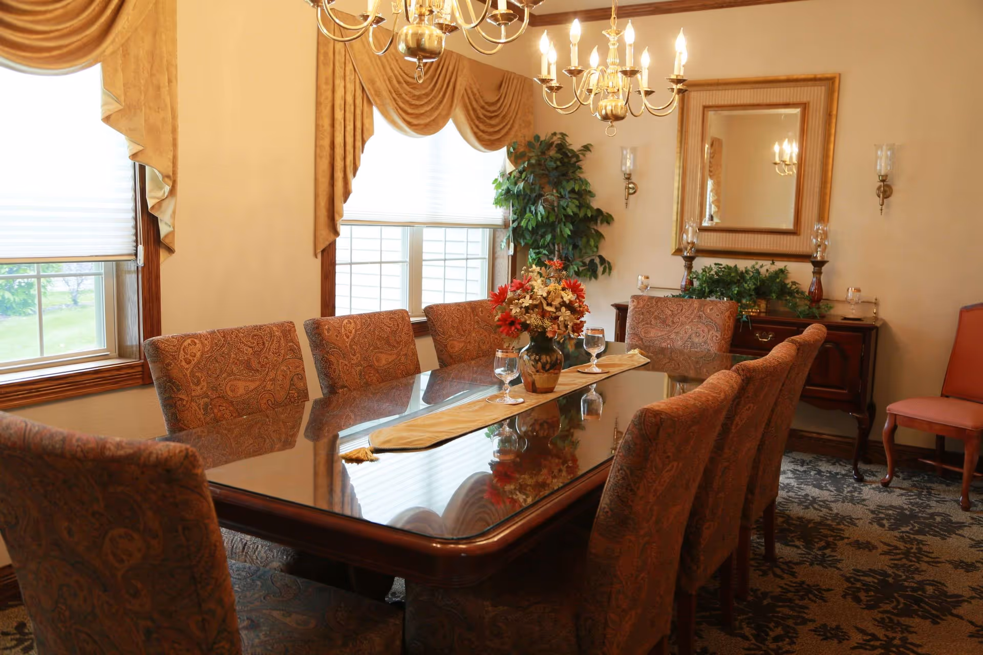 A formal dining room with a large polished wooden table surrounded by eight upholstered chairs with a paisley pattern. The table has a beige runner and a vase with a floral arrangement in the center, along with three water glasses. The room has two windows with white blinds and gold drapes, a chandelier with lit candles hanging above the table, a large mirror on the wall, a wooden sideboard with decorative plants and candle holders, and a patterned carpet.