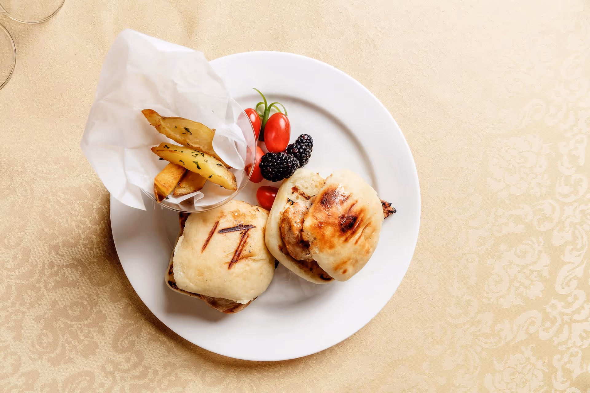 A white plate on a patterned beige tablecloth with two grilled sandwiches, a small serving of potato wedges in a paper cone, and a few cherry tomatoes and blackberries as garnish.
