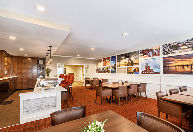 A bright and spacious dining area with multiple wooden tables and brown leather chairs arranged neatly. The room features a red carpet and white walls adorned with colorful framed photographs of landscapes and buildings. On the left side, there is a counter with a buffet setup and red cushioned bar stools. The ceiling has recessed lighting and hanging pendant lights above the counter.