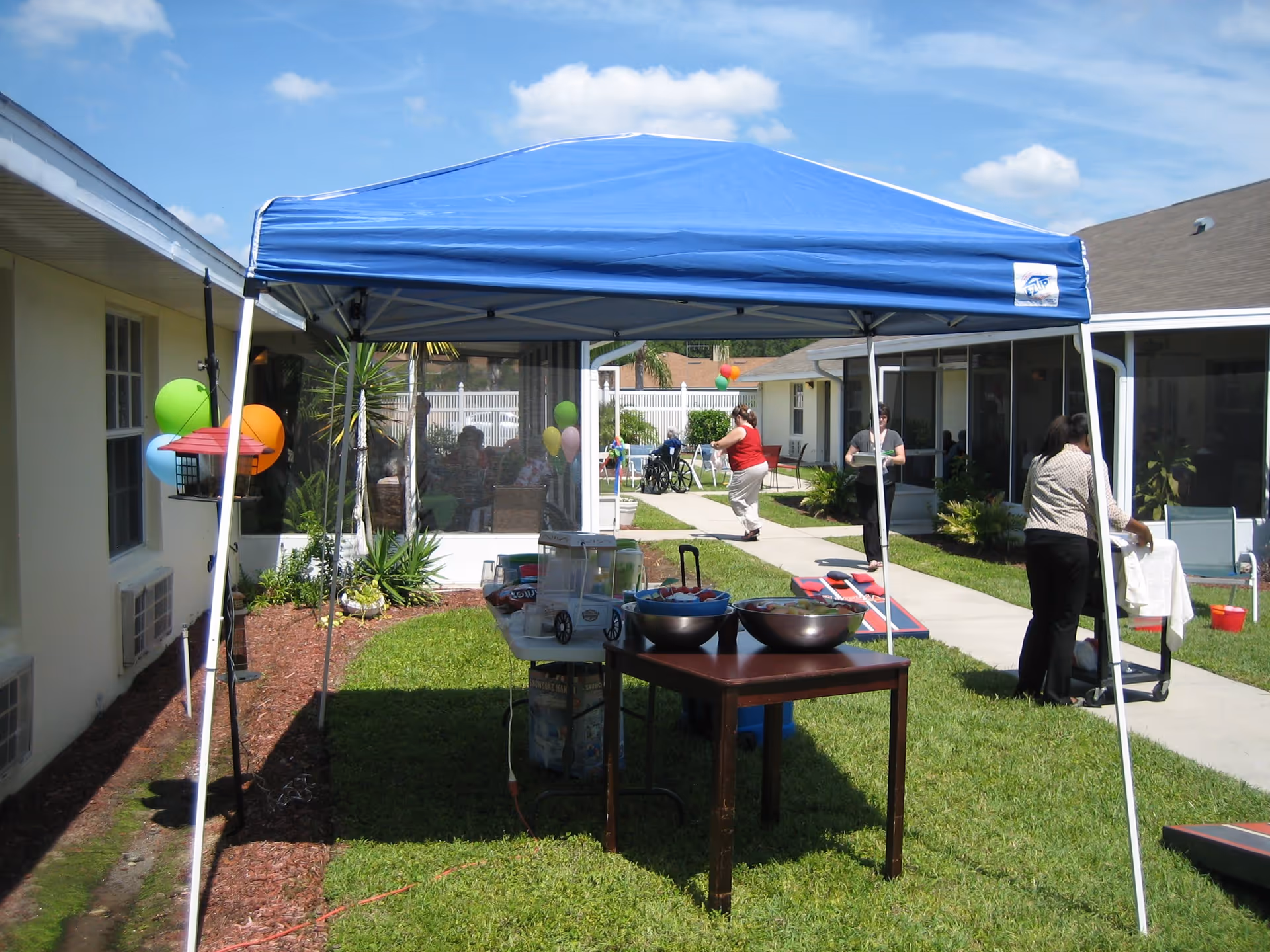 Outdoor scene at Hawthorne Inn with a blue canopy tent set up on a grassy area. Under the tent, there are tables with bowls and containers, likely for a gathering or event. People are walking and standing around the area, with balloons and a cornhole game visible in the background. The setting is sunny with a clear blue sky.