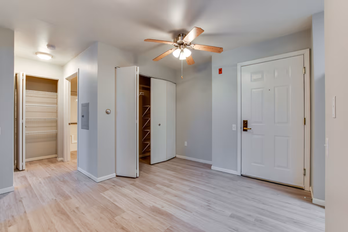 Empty apartment entry and living area with a white front door, open closets, ceiling fan, and light wood floors.