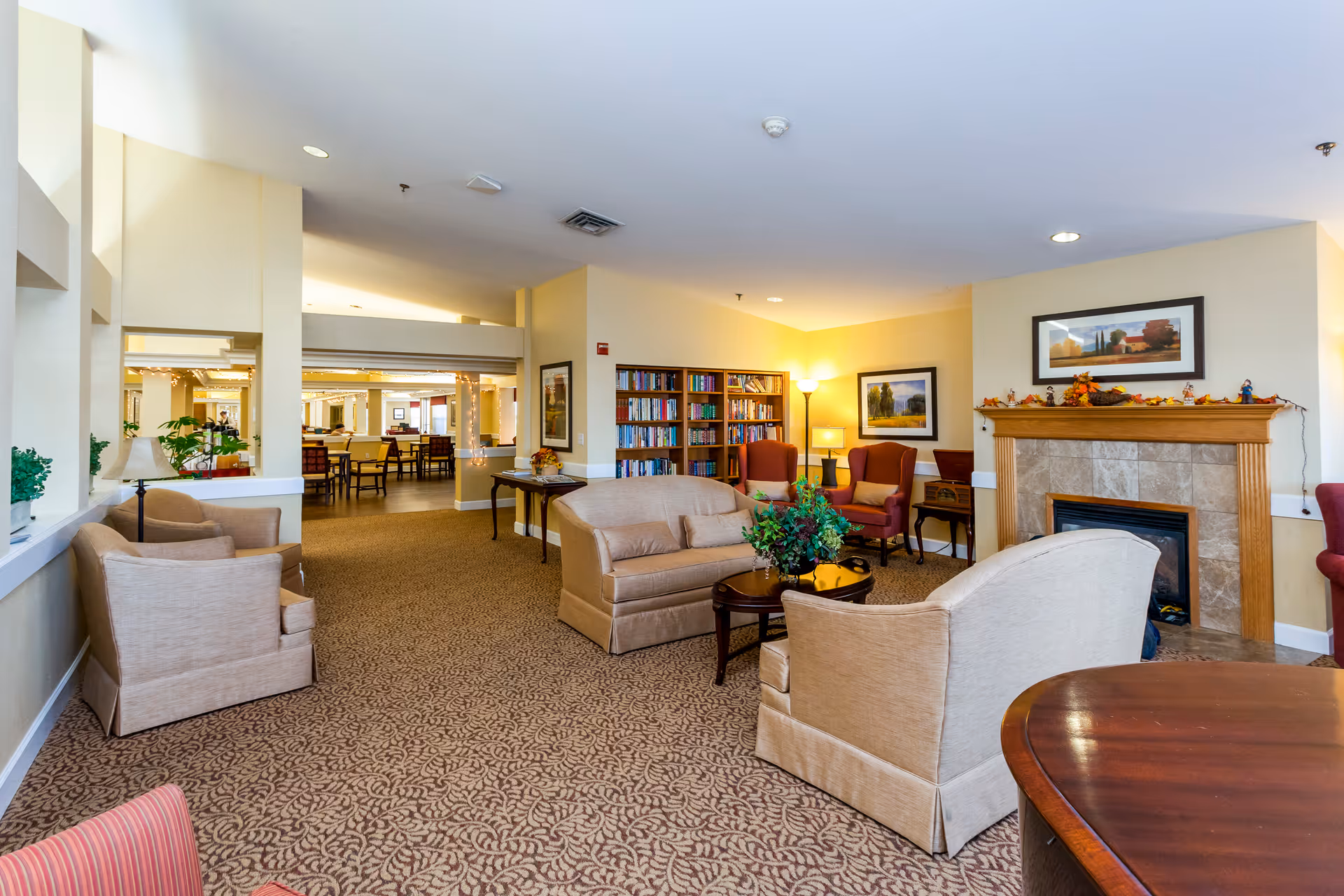 A cozy senior living facility common area with beige upholstered chairs and sofas arranged around a small wooden coffee table with a plant centerpiece. There is a fireplace decorated with autumn-themed ornaments and framed landscape paintings on the walls. A bookshelf filled with books and two red armchairs with pillows are visible in the background. The room is warmly lit with floor and table lamps, and an adjacent dining area with tables and chairs can be seen through an open partition.
