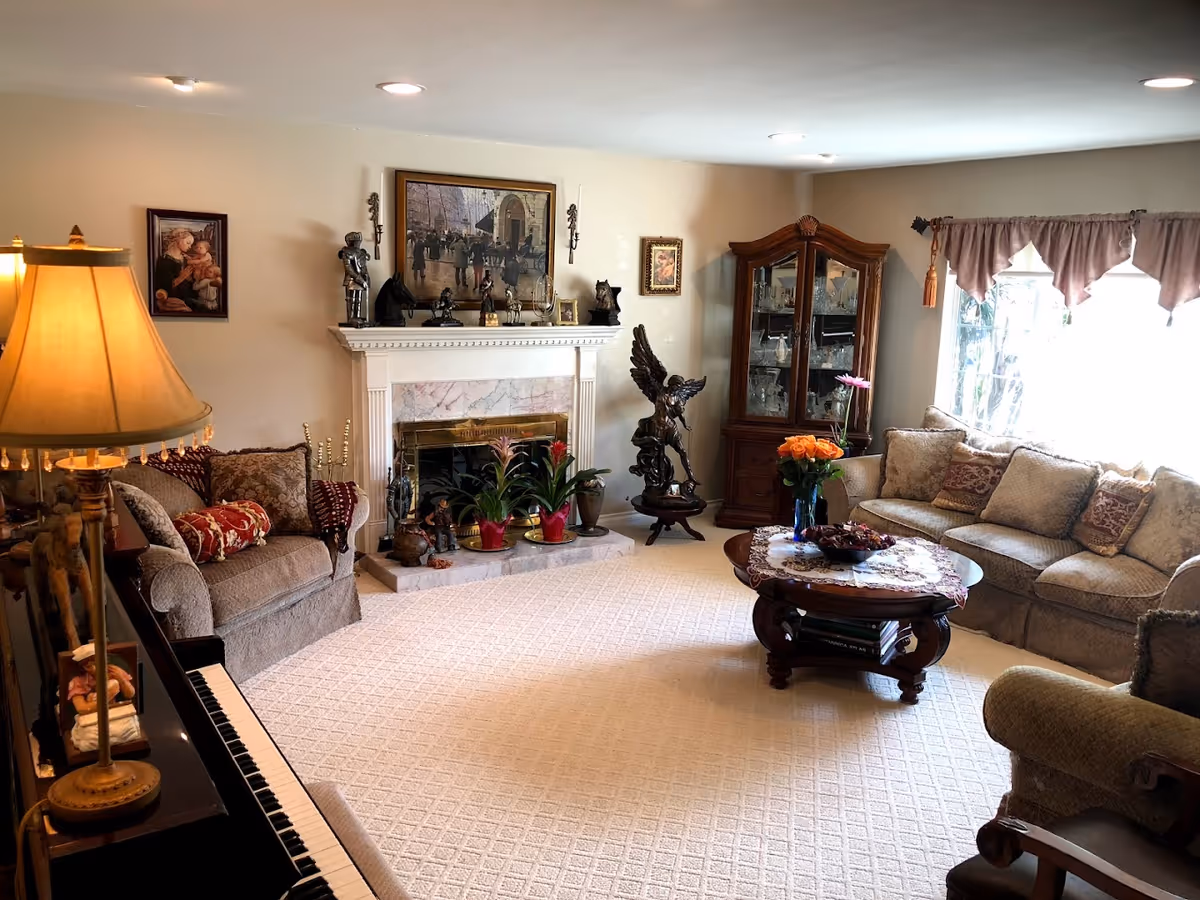 A cozy living room with beige carpet and walls, featuring a fireplace with decorative items on the mantel, a glass-front wooden cabinet filled with glassware, a coffee table with a floral centerpiece, a sofa and armchairs with patterned cushions, a piano with a lamp on it, and a large window with curtains allowing natural light to enter.