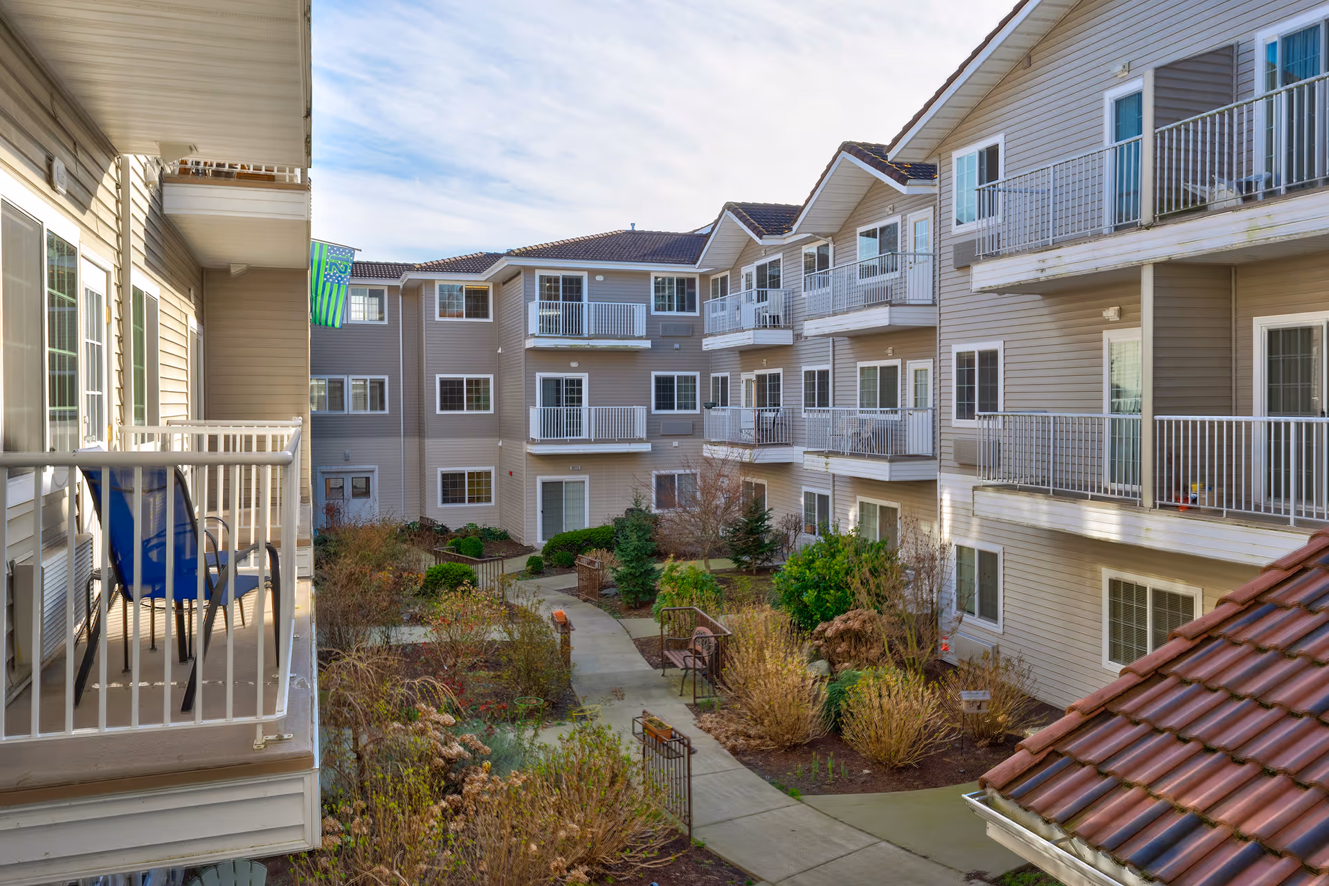 View of a multi-story residential building with balconies overlooking a landscaped courtyard with pathways, shrubs, and small trees under a partly cloudy sky.