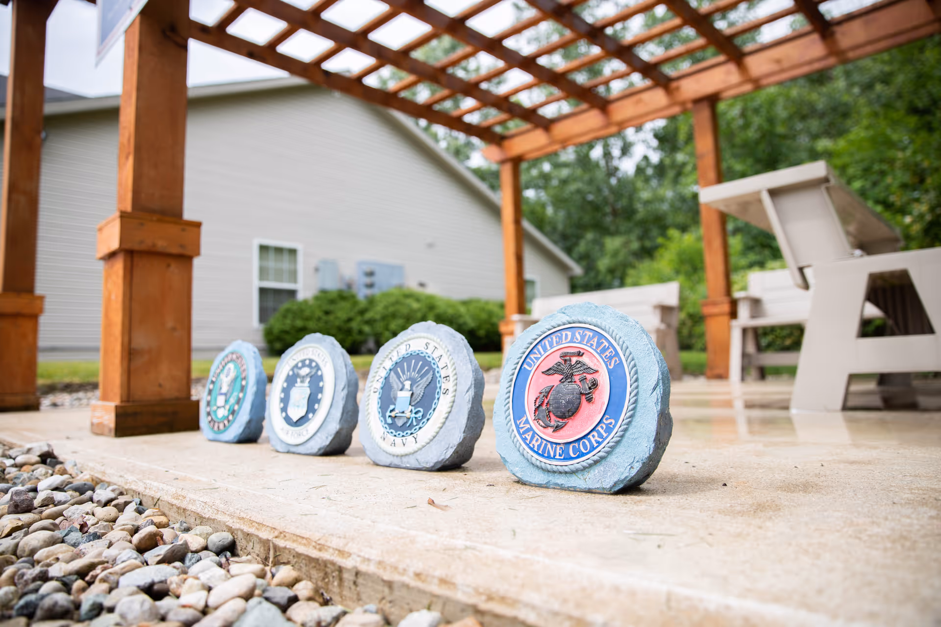 Outdoor patio area with a wooden pergola and white chairs. Four decorative stones with emblems of United States military branches including Marine Corps, Navy, Air Force, and Army are lined up on the concrete floor. A building and greenery are visible in the background.