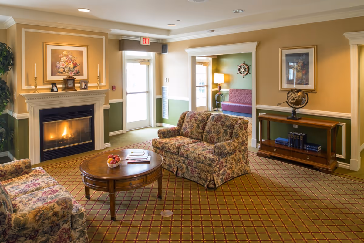 A cozy living room area in an assisted living facility featuring floral upholstered sofas, a wooden coffee table with a bowl of fruit and books, a lit fireplace with decorative candlesticks and framed artwork above it, a wooden console table with books and a globe, and an exit door letting in natural light.
