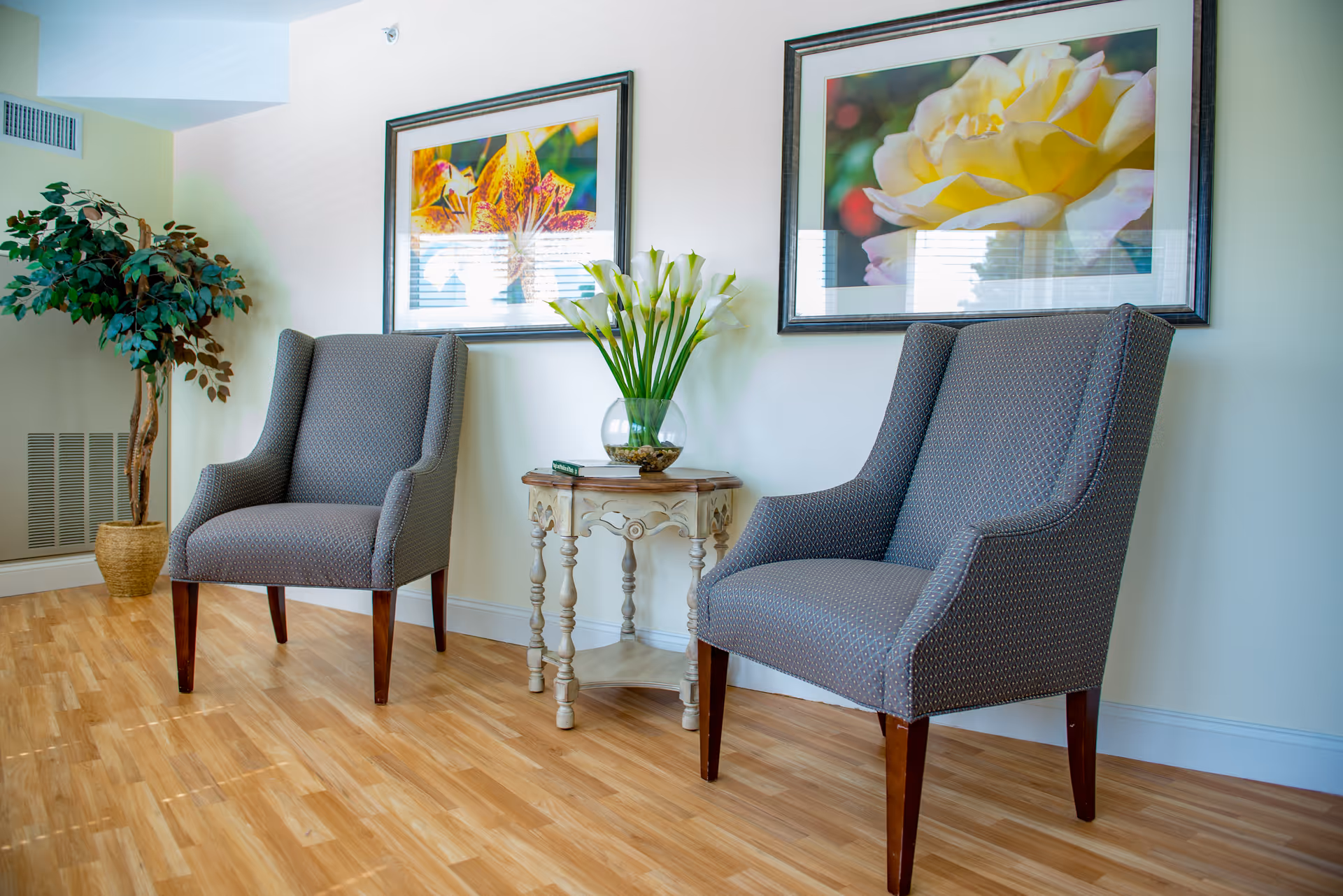 A cozy seating area with two gray upholstered armchairs placed on a wooden floor. Between the chairs is a small ornate wooden table holding a glass vase with white calla lilies. Behind the chairs, two framed floral photographs hang on a light-colored wall. A potted artificial tree is positioned in the corner of the room.