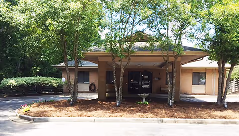 Brick facility entrance with glass doors, shaded by trees and a small driveway.