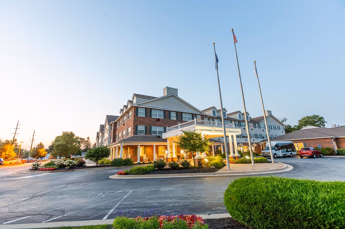 Exterior view of a multi-story senior living facility building with brick and siding facade, a covered entrance with white columns, three flagpoles, landscaped bushes and flowers, parked cars, and a clear sky at dusk.