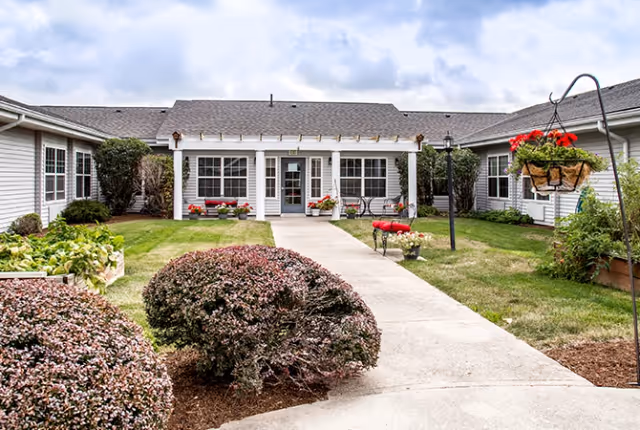 A well-maintained courtyard with a central walkway leading to a single-story building entrance, surrounded by shrubs, hanging flower baskets, and benches.