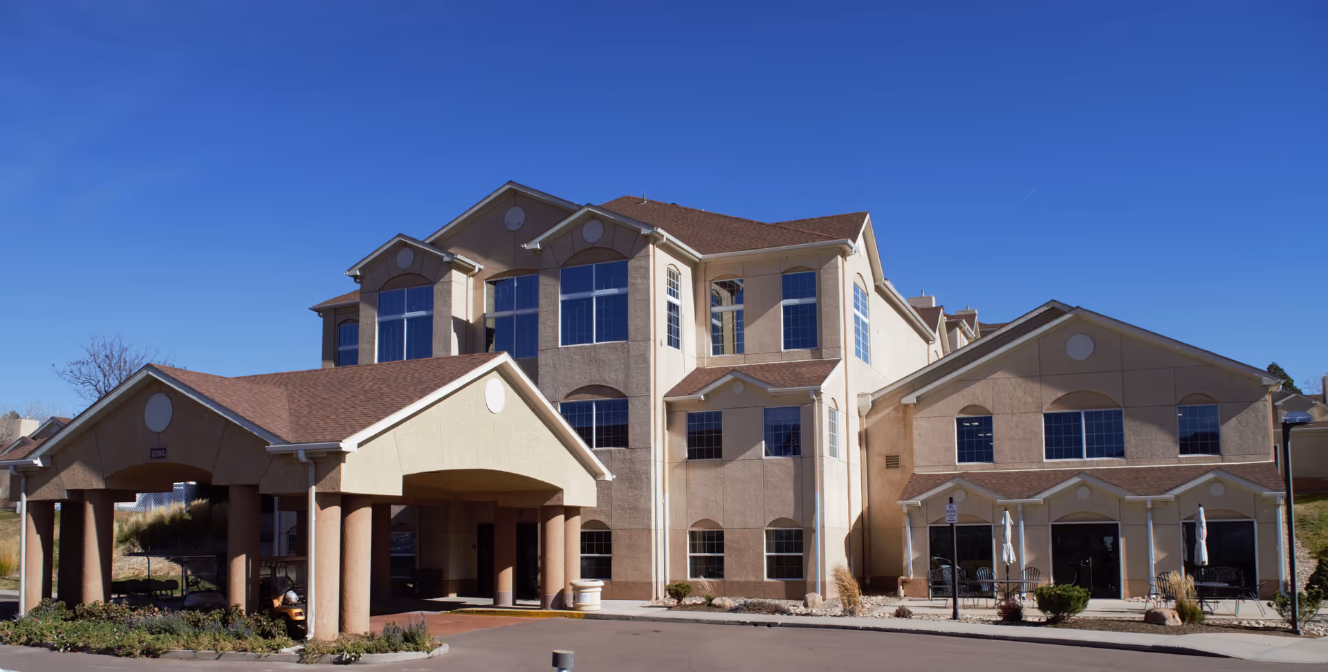 Exterior view of a multi-story senior living facility building under a clear blue sky. The building has beige walls, multiple large windows, and a covered entrance supported by columns. There are outdoor seating areas with tables and chairs near the entrance.