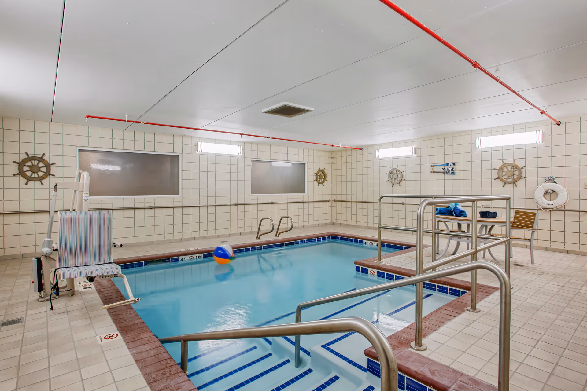 Indoor swimming pool with tiled walls and floor, metal handrails, a pool lift chair, a beach ball floating in the water, and nautical-themed decorations including ship wheels and a life preserver. There is a small table with towels and chairs on the pool deck.