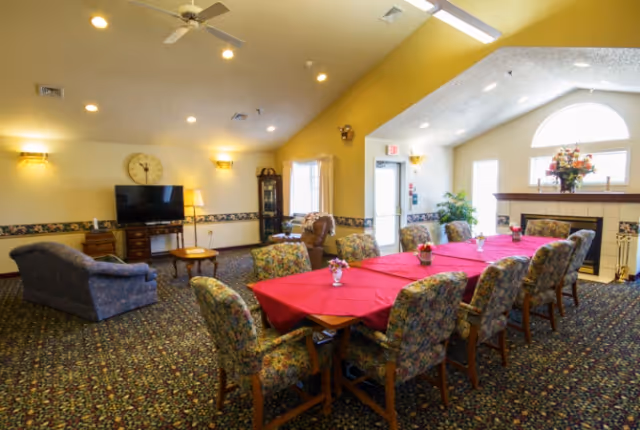 A senior living facility common area with a long table covered in a red tablecloth surrounded by floral upholstered chairs. The room has a patterned carpet, a fireplace with a floral arrangement on the mantel, a TV on a wooden stand, a grandfather clock, and several armchairs. The ceiling is vaulted with recessed lighting and a ceiling fan.