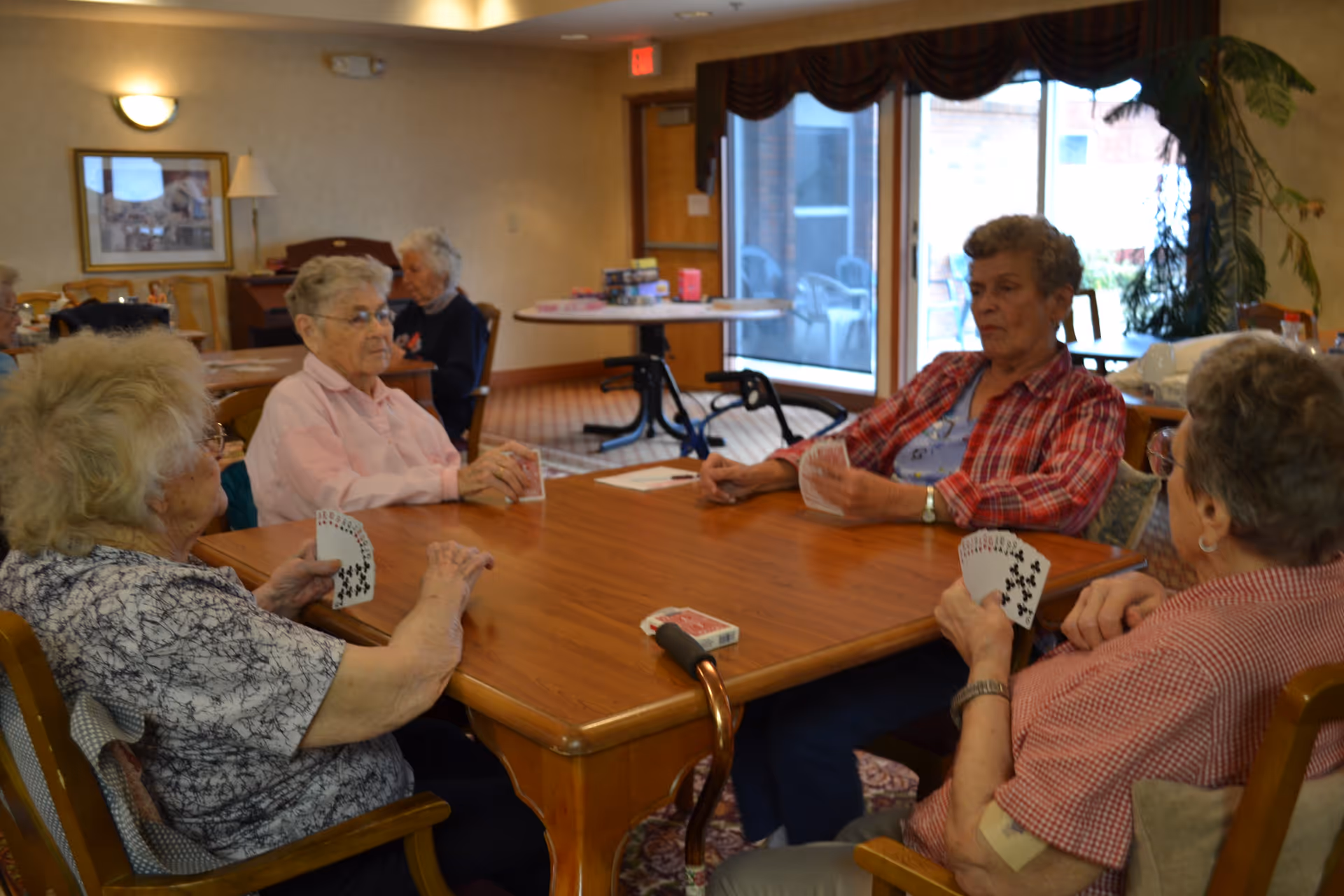 Four elderly women sitting around a wooden table playing cards in a well-lit room with large windows and a sliding glass door. The room has carpeted floors, a plant near the window, and a few tables and chairs in the background.