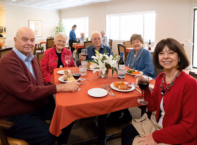 Five older adults seated around a table in a dining room, smiling and enjoying a meal and glasses of wine.