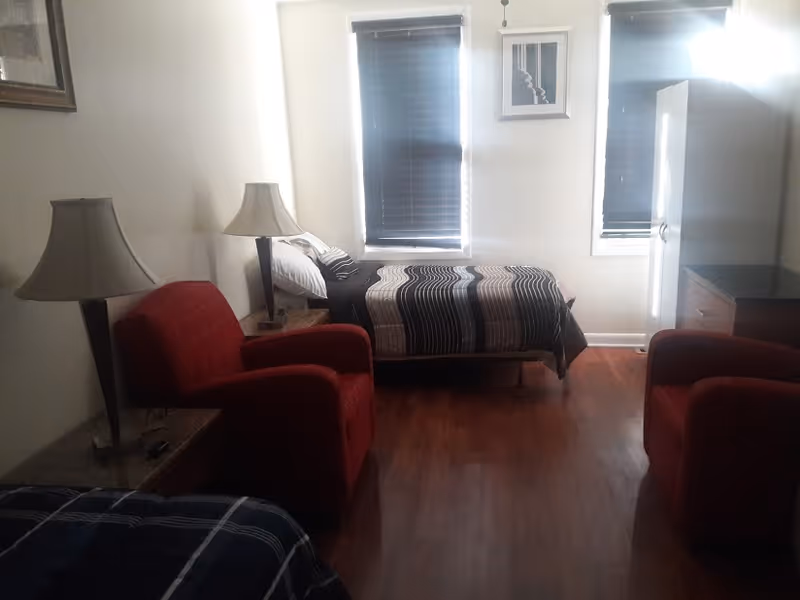 Small furnished bedroom with two twin beds, two red armchairs, bedside lamps, a wardrobe, and two windows with blinds.