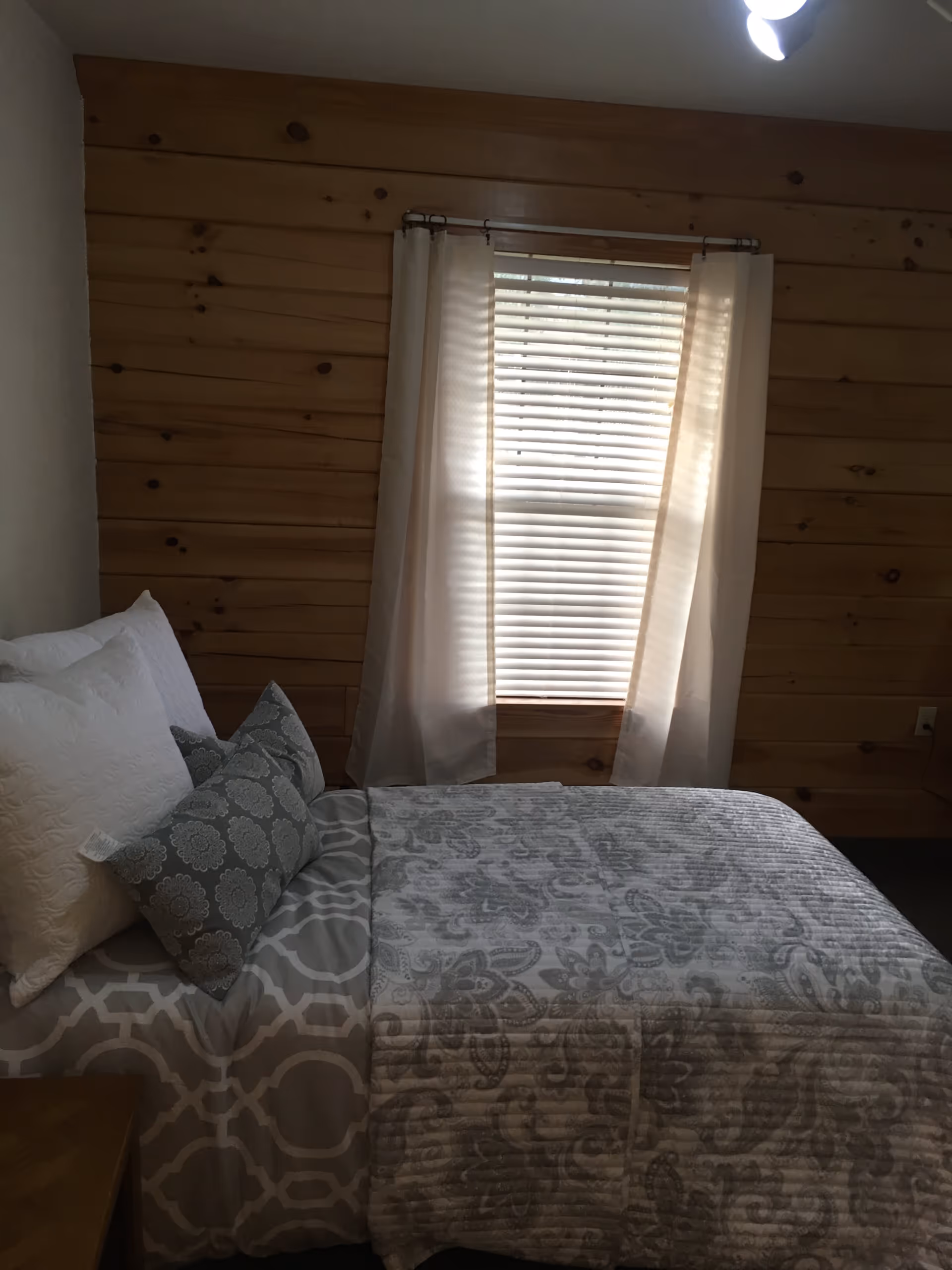 Cozy bedroom with a made bed, patterned bedding and pillows, a window with blinds and curtains, and wood-paneled walls.