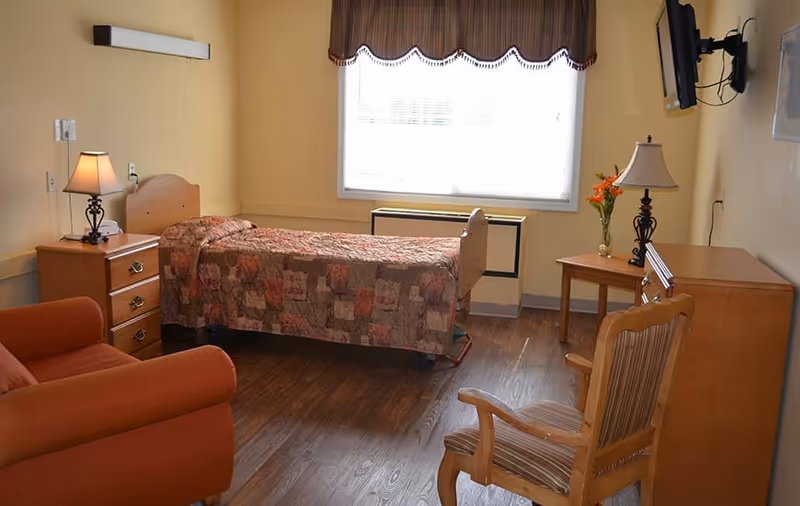 A furnished senior bedroom with a single bed, nightstand, armchair, wooden chair, dresser, lamps and a wall-mounted TV beneath a bright window.