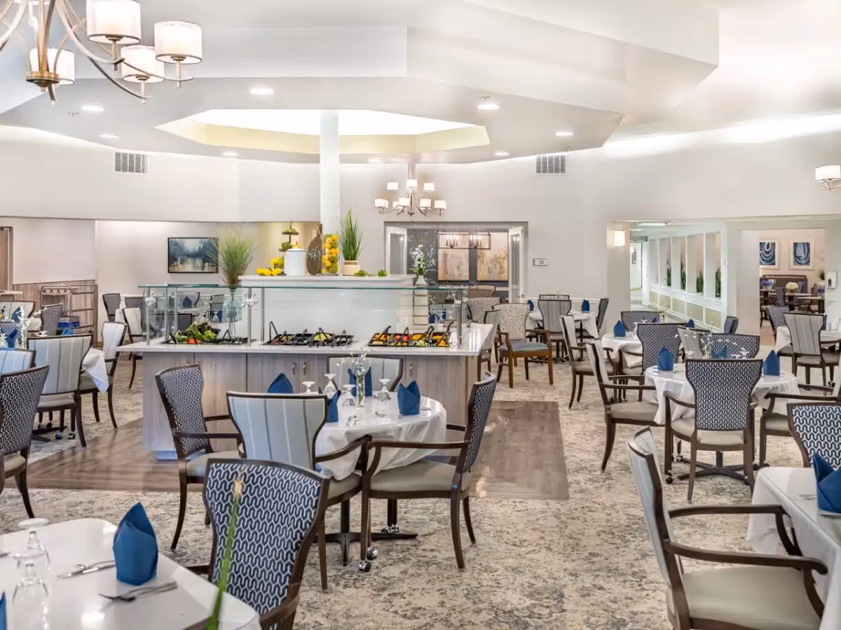 A spacious and well-lit dining room with multiple round tables covered with white tablecloths and set with blue napkins, glassware, and silverware. The room features patterned chairs, a central buffet station with various food items, decorative plants, and modern chandeliers hanging from the ceiling.
