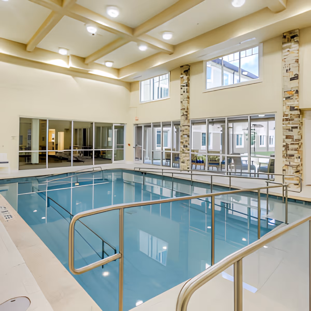 Indoor swimming pool with handrails and steps leading into the water, surrounded by large windows and beige walls with stone accents, located inside a senior living facility.