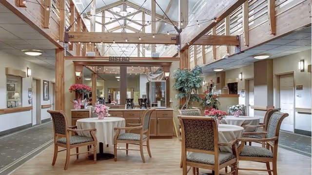 A bright and spacious dining area in a senior living facility with round tables covered in white tablecloths, surrounded by wooden chairs with patterned cushions. The room features a high ceiling with exposed wooden beams and skylights, allowing natural light to fill the space. There are decorative plants and flowers on the tables and around the room, and a sign reading 'BISTRO' above a counter area in the background.