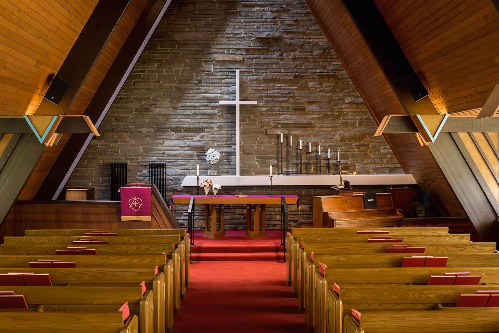 Interior view of a chapel with wooden pews, a red carpet aisle, a wooden altar covered with a purple cloth, a white cross mounted on a stone wall, and a piano on the right side.