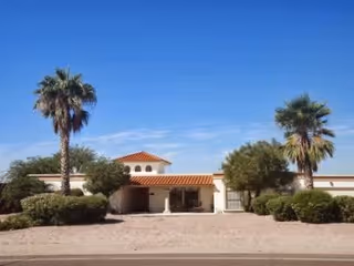 Single-story stucco assisted living building with a tiled roof and palm trees in front under a clear blue sky.