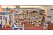 Interior view of a small pantry or food storage room with shelves stocked with various canned and packaged food items. There is a refrigerator on the left side and a counter with additional items in the foreground. The room is well-lit with fluorescent lighting.