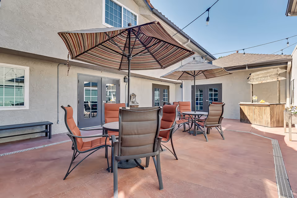 Sunlit outdoor courtyard patio with tables, striped umbrellas, and cushioned chairs outside a senior living building.