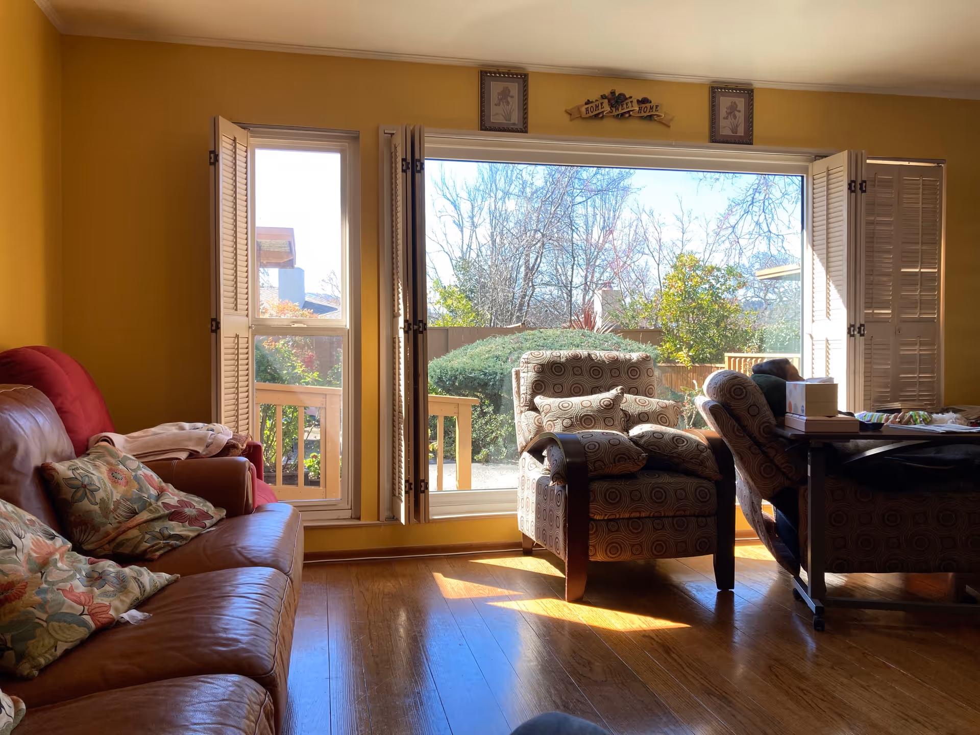 A cozy living room with a large window and glass door opening to a garden outside. The room has yellow walls, wooden flooring, a brown leather sofa with floral cushions, and two patterned armchairs. Sunlight streams through the window, illuminating the space. Above the window is a decorative sign that reads 'Home Sweet Home'.
