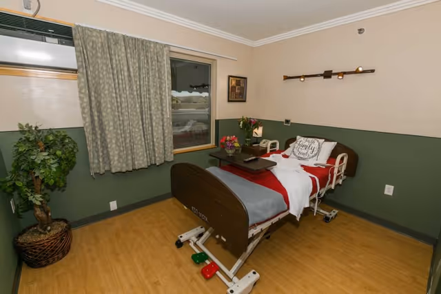 A small, clean patient room with a hospital bed made up with white, red, and gray bedding. A pillow with the words 'Family is everything' is on the bed. There is a wooden tray table on the bed with a TV remote. A potted plant is in the corner near a window with closed curtains. The walls are painted half green and half beige, and there is a wall-mounted light fixture above the bed.
