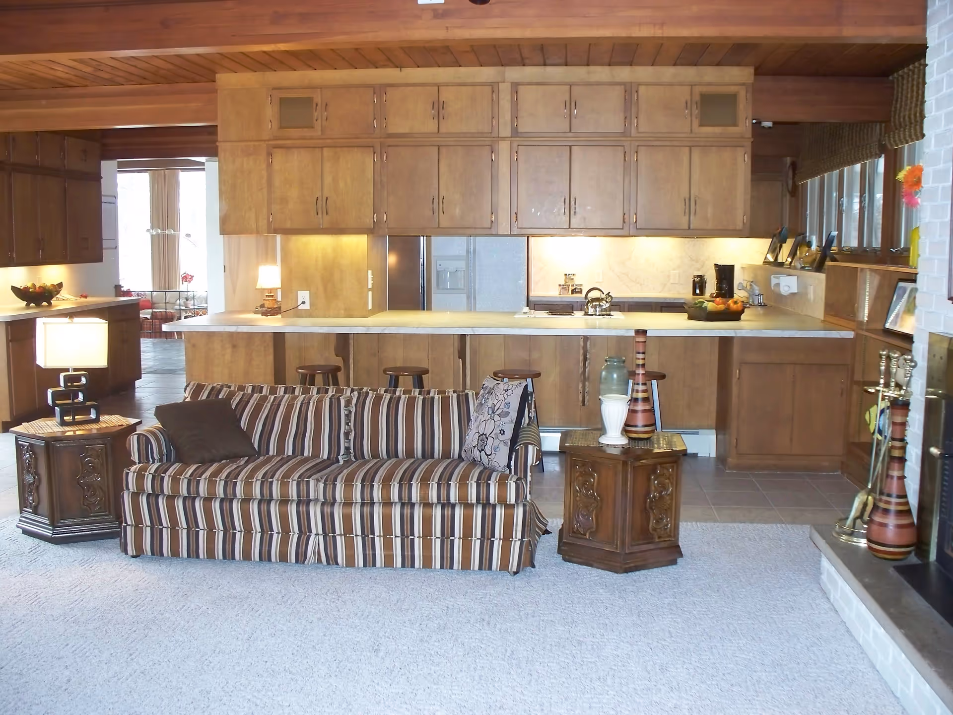 Interior view of a living room and kitchen area in The Sugarbush House. The living room features a striped sofa with two pillows, a wooden side table with a lamp, and another wooden table with decorative vases. The kitchen has wooden cabinets, a countertop with three stools, a refrigerator, a coffee maker, and a bowl of fruit. The room has a carpeted floor and a fireplace with decorative items on the right side.