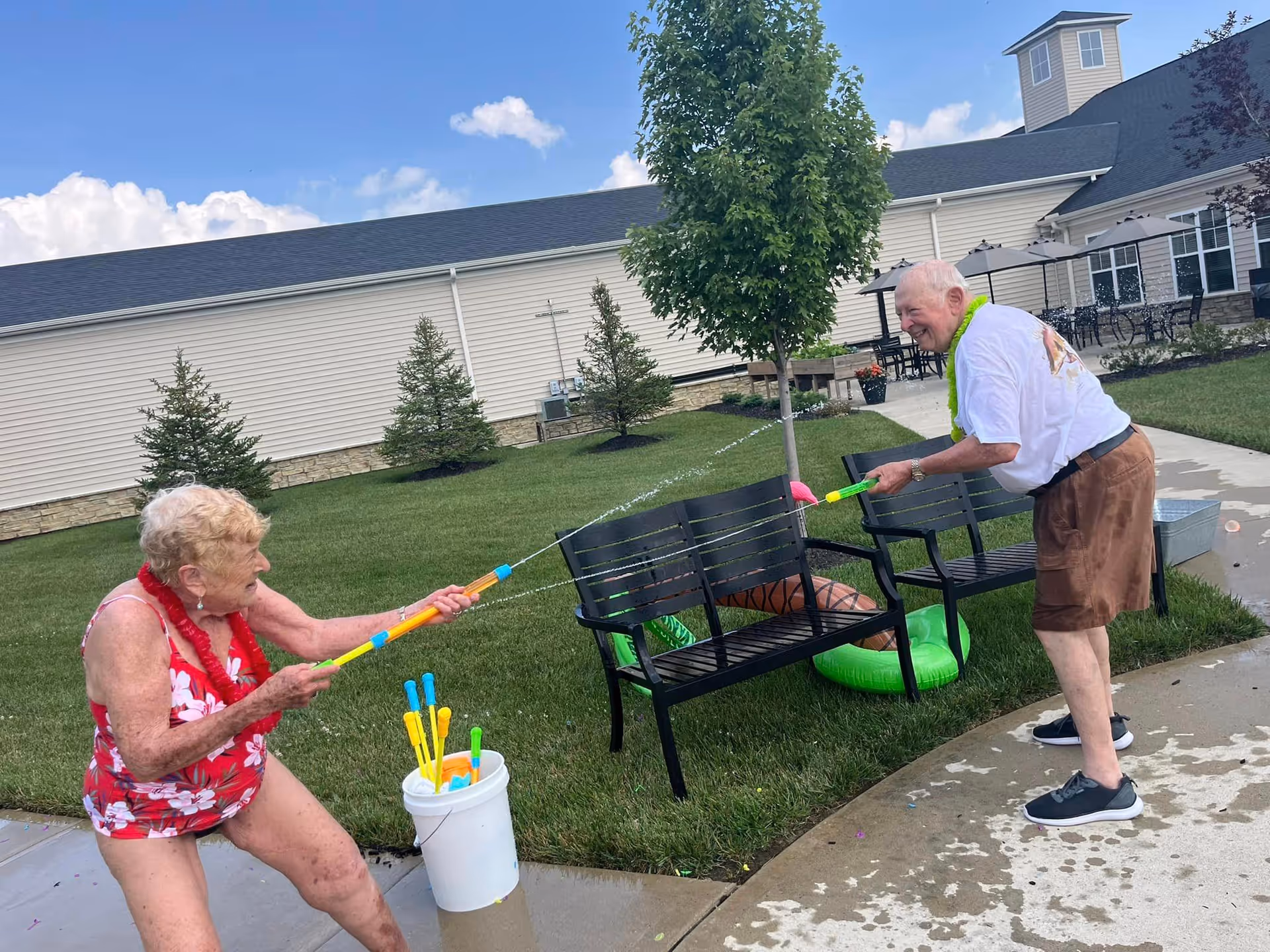Two elderly people having fun outdoors playing with water guns near a bench and green lawn, with a building and trees in the background under a blue sky with some clouds.