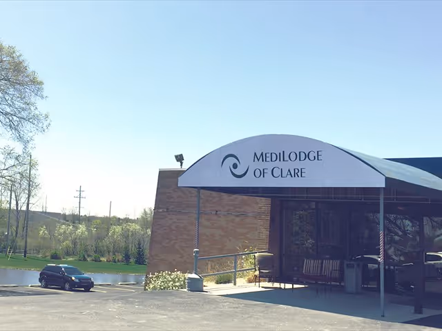 Exterior view of Medilodge of Clare showing the entrance with a covered awning displaying the facility's name. There is a parking lot with a few cars and some greenery in the background.
