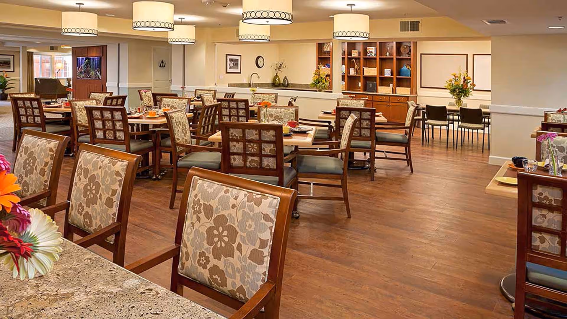 A spacious dining room with multiple wooden tables and chairs featuring floral upholstery. The room has warm wooden flooring, modern ceiling lights, and decorative flowers on the tables. In the background, there is a built-in wooden shelving unit and a small counter area with a sink.