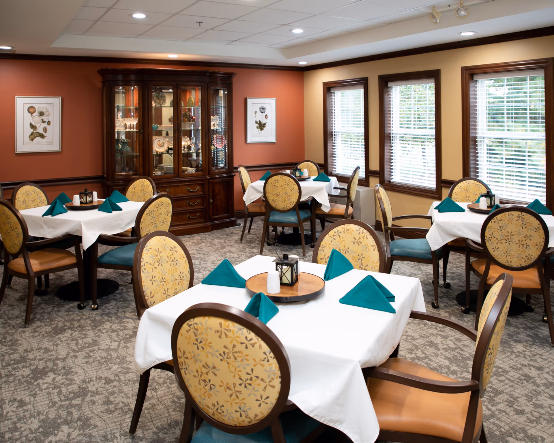 A dining room with several square tables covered with white tablecloths and set with green folded napkins. Each table has four chairs with patterned yellow and blue upholstery. There is a wooden china cabinet against a reddish-brown wall displaying glassware and decorative plates. Three windows with white blinds let in natural light on the right side of the room. The carpet has a subtle floral pattern.